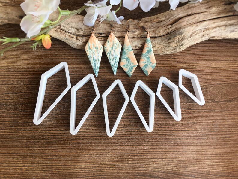 May include: Four diamond-shaped earrings with a marbled peach and teal pattern on copper-toned hooks, accompanied by six white plastic diamond-shaped cutters of varying sizes arranged in a row below the jewelry pieces.