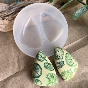 May include: A clear silicone mold with two teardrop-shaped cavities, alongside two finished leaf-patterned pendants. The pendants are light yellow with detailed green leaf designs. The mold is for crafting resin jewelry.