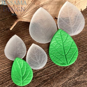 May include: A collection of leaf-shaped silicone molds in various sizes, including two opaque green leaf-shaped pieces and four translucent white leaf-shaped molds, all featuring detailed vein patterns on their surfaces for crafting.