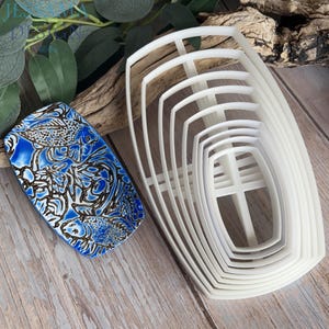 May include: A rectangular tray featuring a blue and white fish pattern alongside a set of seven nested white plastic cookie cutters. The tray displays a detailed illustration of multiple fish with dark outlines, while the cutters are shaped like rounded pentagons of varying sizes, stacked together to show their graduated dimensions.