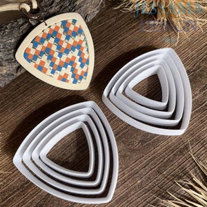 May include: A collection of white, triangular cookie cutters in graduated sizes, alongside a pendant with a woven pattern in blue, orange, and gold. The pendant is attached to a tan leather cord. The items are arranged on a wooden surface.
