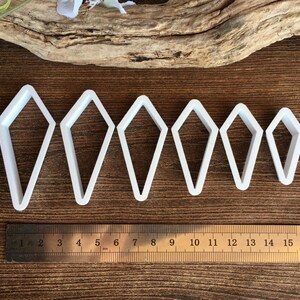 May include: A set of six white kite-shaped cutters arranged in descending order of size. A stainless steel ruler is positioned below the cutters, showing the largest cutter is approximately 3.5 inches tall and the smallest is 2 inches tall.