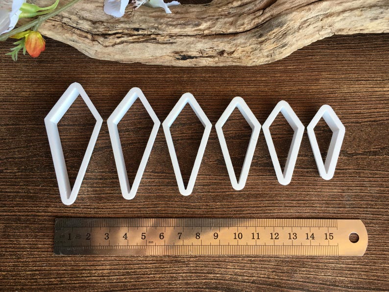 May include: A set of six white kite-shaped cutters arranged in descending order of size. A stainless steel ruler is positioned below the cutters, showing the largest cutter is approximately 3.5 inches tall and the smallest is 2 inches tall.
