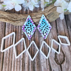 Op de afbeelding: Ruitvormige oorbellen met een blauw en zwart stippenontwerp, omlijst door paarse en witte accenten. Witte ruitvormige uitsteekvormen zijn ook aanwezig. De oorbellen en uitsteekvormen worden gepresenteerd op een houten oppervlak met bloemaccenten.