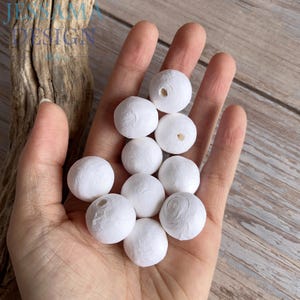 May include: A collection of white, spherical beads with a textured surface and a hole through the center. The beads are held in an open hand, with a wooden background. The beads are approximately 0.75 inches in diameter.