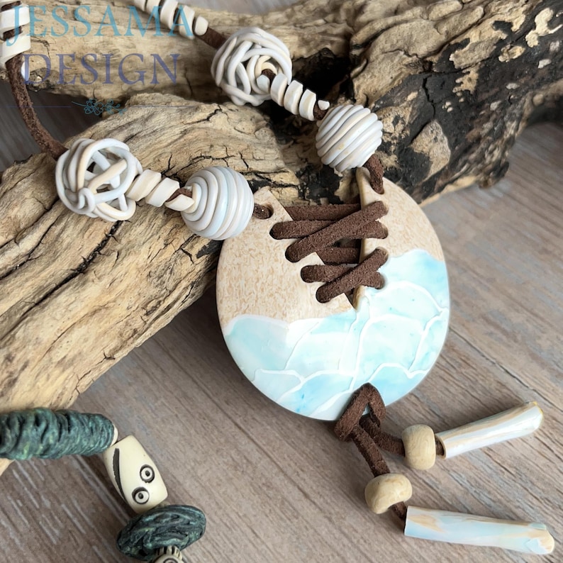 May include: A handcrafted necklace featuring a large, round pendant with a two-tone design of beige and blue, resembling a seascape. The necklace includes various white and blue beads and a brown cord. The text "ESSAM DESIGN" is visible.