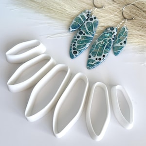 May include: Earrings with a teal and white design, featuring a butterfly wing pattern, are displayed next to white oval-shaped clay cutters. The earrings have silver hooks. The background includes a white surface and a light-colored, feathery accent.