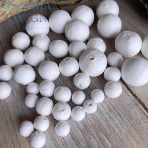 May include: A collection of white, spherical beads of varying sizes. Some beads have small holes. The beads are arranged on a light brown, wooden surface. The image is well-lit, highlighting the texture of the beads.