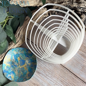 May include: A set of white plastic circular cookie cutters nested together, featuring a graduated size design. Beside the cutters is a small, round, textured dish with a vibrant blue and gold abstract pattern across its surface.
