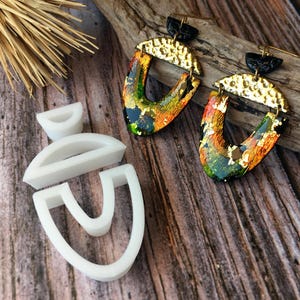 May include: A pair of colorful, abstract earrings with gold accents. The earrings are made of polymer clay and have a unique, textured finish. The earrings are hanging from a wooden surface.