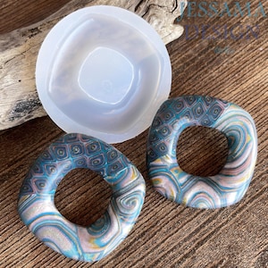 May include: A translucent silicone mold and two square-shaped charms with rounded corners. The charms feature a multi-colored pattern of teal, blue, and metallic silver swirls and geometric grid designs with a smooth, glossy finish.