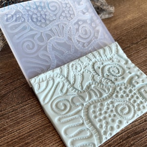 Pode incluir: Um molde de silicone azul claro com um intrincado padrão floral e de redemoinhos em relevo. Uma telha de argila azul clara correspondente é parcialmente removida do molde, mostrando o design detalhado. A palavra "JESSAMA DESIGN" é visível.