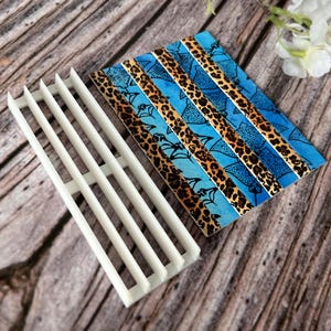 Könnte beinhalten: Ein weißer Seifenschneider aus Kunststoff neben einem dekorativen Seifenstück. Das Seifenstück hat ein blaues und Leopardenmuster. Der Schneider hat eine Reihe paralleler Stäbe. Die Artikel befinden sich auf einer Holzoberfläche.