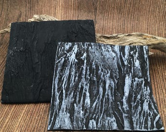 May include: Two square coasters with a textured surface. One coaster is solid matte black, while the other features a black base with silver highlights applied to the raised, uneven ridges, creating a high-contrast, weathered appearance.