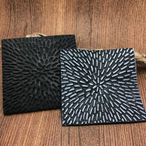 May include: Two square coasters with a raised, textured design. One coaster is black and the other is a dark gray with a silver sheen.