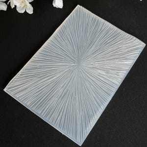 Könnte beinhalten: Eine rechteckige, durchscheinende Silikonform mit einem strukturierten, radialen Design. Das Muster ähnelt Linien, die von einem zentralen Punkt ausgehen und einen Sonnenstrahleffekt erzeugen. Die Form ist wahrscheinlich für Bastel- oder Harzprojekte gedacht.