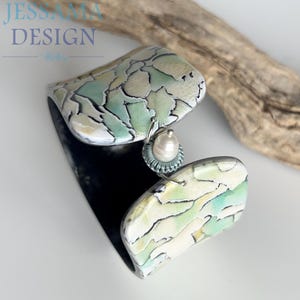 May include: A cuff bracelet featuring a cracked pattern in shades of mint green, cream, and black. The two ends are connected by a silver-toned metal link holding a single white freshwater pearl surrounded by a small, textured teal-coloured ring.