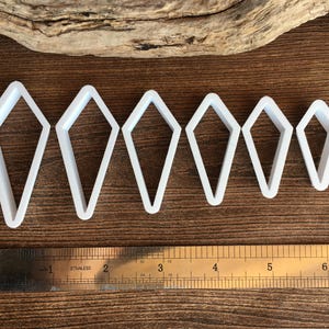 May include: Six white kite-shaped cutters arranged in a row from largest to smallest. A stainless steel ruler is positioned below the cutters, showing the largest cutter is approximately 2.5 inches tall and the smallest is 1.5 inches tall.