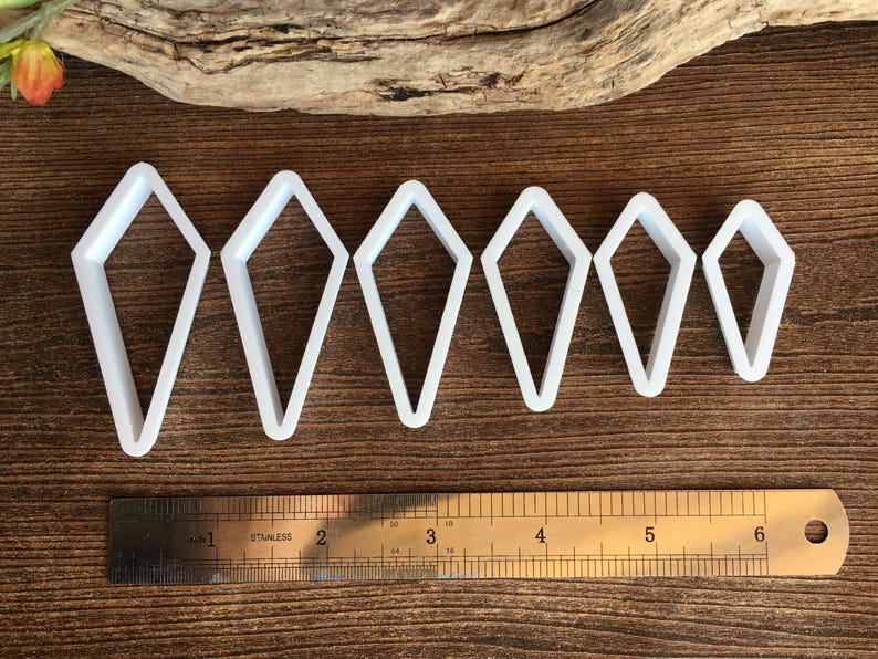 May include: Six white kite-shaped cutters arranged in a row from largest to smallest. A stainless steel ruler is positioned below the cutters, showing the largest cutter is approximately 2.5 inches tall and the smallest is 1.5 inches tall.