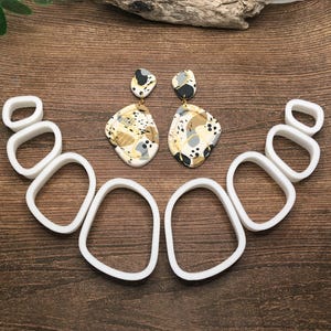 May include: A set of six white plastic cookie cutters in various sizes and shapes, resembling pebbles or stones. The cutters are arranged in a semi-circle on a wooden surface. Two pairs of earrings with abstract patterns in shades of gray, black, and gold are also shown on the surface.