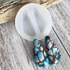 Pode incluir: Um molde de silicone redondo e translúcido com duas cavidades em forma de lágrima, acompanhado por dois pingentes acabados em forma de lágrima com um padrão de mosaico de segmentos azul-claros, brancos e cor de cobre metálico.