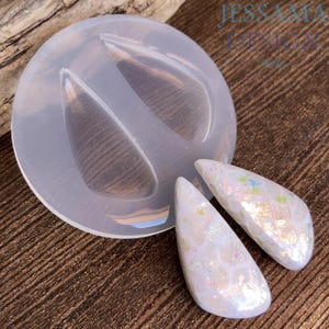 May include: A clear silicone mold with two teardrop-shaped cavities, next to two finished white and iridescent teardrop-shaped resin pendants. The pendants have a textured surface and reflect multiple colors. The text "JESSAMA DESIGN" is visible in the upper right corner.