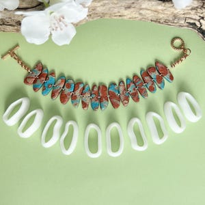 May include: A handmade bracelet with a toggle clasp, featuring oval-shaped beads in shades of red, blue, and brown. The bracelet is displayed with several white oval-shaped craft tools, all set against a light green background.