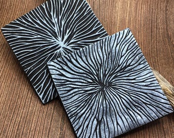 May include: Two square black coasters with a white, raised, radial pattern. The coasters are made of a textured material and have a slightly rough surface.