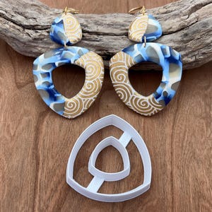 May include: A pair of blue and gold statement earrings with a geometric design. The earrings are made of polymer clay and have a unique, abstract pattern. The earrings are shown with a white plastic mold.
