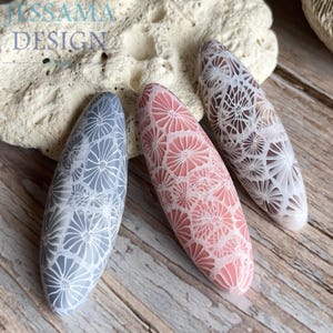 Può includere: Tre design ovali per nail art nei toni del grigio, rosa e marrone, ciascuno con un delicato motivo floreale. I design sono esposti su una superficie di legno. Il testo "JISSAMA DESIGN" è visibile in alto a sinistra.