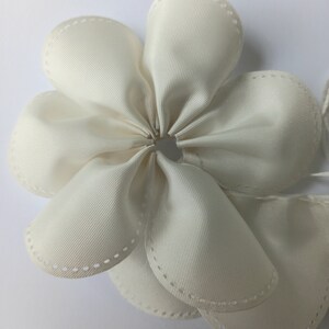 May include: A white fabric flower with a central hole. The flower is made of multiple layers of fabric and has a stitched edge.