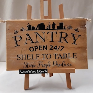 May include: Wooden sign with black text that reads "Pantry Open 24/7 Shelf to Table Store Fresh Produce" with a small silhouette of a kitchen above the word "Pantry". The sign is on a small wooden easel with the text "Aussie Wood & Crafts" at the bottom.