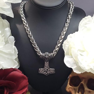 May include: A silver chain necklace with a silver Mjolnir pendant. The pendant is a detailed design with a knotwork pattern. The necklace is displayed on a black mannequin with white flowers and a skull on either side.