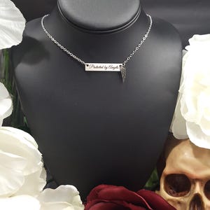 K&ouml;nnte beinhalten: Eine silberfarbene Halskette mit einem rechteckigen Anh&auml;nger mit der Aufschrift "Protected by Angels". Ein kleiner Fl&uuml;gelanh&auml;nger h&auml;ngt daneben. Die Halskette wird auf einer schwarzen B&uuml;ste pr&auml;sentiert, umgeben von wei&szlig;en Blumen, roten Rosen und einem Totenkopf.