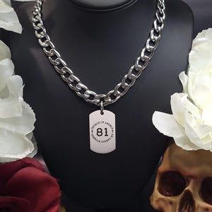 Peut inclure: Un collier en chaîne argentée avec un pendentif en forme de plaque d'identité argentée. La plaque d'identité porte le texte "Church of Angels + Suffolk County, NY +" et le numéro "81" au centre.