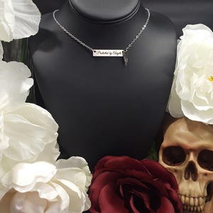 K&ouml;nnte beinhalten: Eine silberne Halskette mit einem rechteckigen Anh&auml;nger mit der Aufschrift "Protected by Angels". Die Halskette wird auf einer schwarzen B&uuml;ste pr&auml;sentiert, umgeben von wei&szlig;en und burgunderfarbenen Blumen und einem Totenkopf.