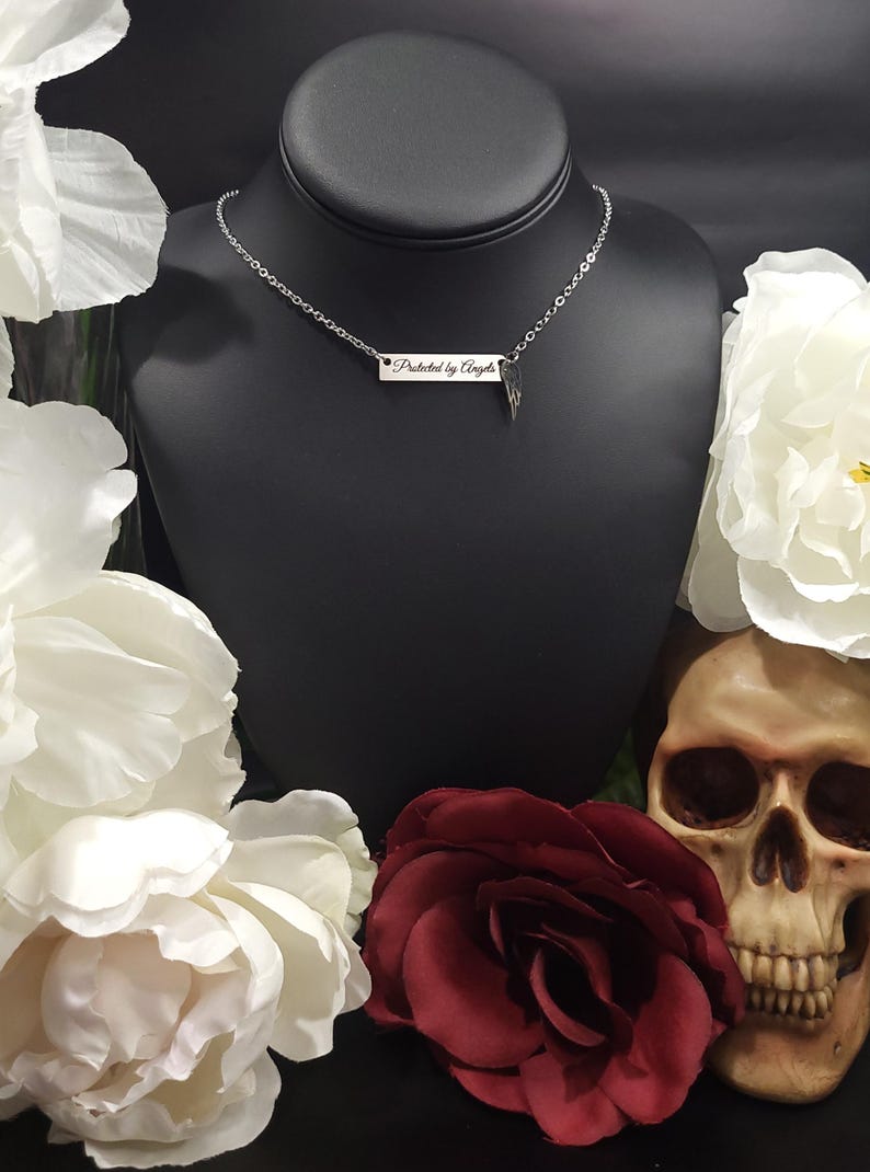 K&ouml;nnte beinhalten: Eine silberne Halskette mit einem rechteckigen Anh&auml;nger mit der Aufschrift "Protected by Angels". Die Halskette wird auf einer schwarzen B&uuml;ste pr&auml;sentiert, umgeben von wei&szlig;en und burgunderfarbenen Blumen und einem Totenkopf.