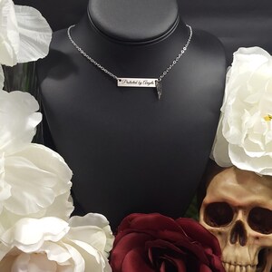 K&ouml;nnte beinhalten: Eine silberne Halskette mit einem rechteckigen Anh&auml;nger mit der Aufschrift "Protected by Angels" und einem kleinen Fl&uuml;gel-Charm. Die Halskette wird auf einem schwarzen Mannequinkopf pr&auml;sentiert, umgeben von wei&szlig;en Blumen, einer roten Rose und einem Totenkopf.