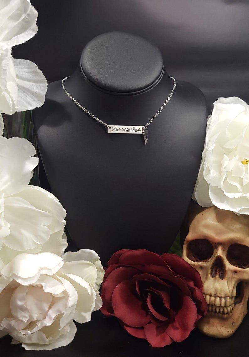 K&ouml;nnte beinhalten: Eine silberne Halskette mit einem rechteckigen Anh&auml;nger mit der Aufschrift "Protected by Angels" und einem kleinen Fl&uuml;gel-Charm. Die Halskette wird auf einem schwarzen Mannequinkopf pr&auml;sentiert, umgeben von wei&szlig;en Blumen, einer roten Rose und einem Totenkopf.