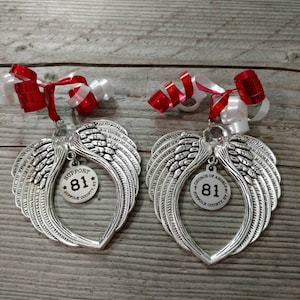 Peut inclure: Deux ornements en forme d'ailes d'ange argentés avec une étiquette ronde qui dit "Support 81 Suffolk County" et "Church of Angels 81 Suffolk County". Les ornements sont suspendus à un ruban rouge et blanc.