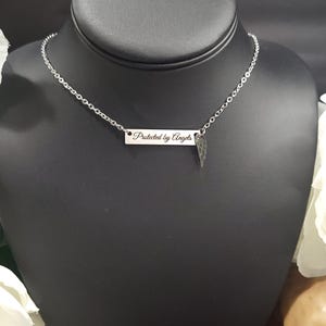 K&ouml;nnte beinhalten: Eine silberfarbene Halskette mit Kette und rechteckigem Anh&auml;nger. Der Anh&auml;nger ist mit den Worten "Protected by Angels" graviert und hat einen kleinen Fl&uuml;gelanh&auml;nger. Die Halskette wird auf einer schwarzen Schaufensterpuppe pr&auml;sentiert.