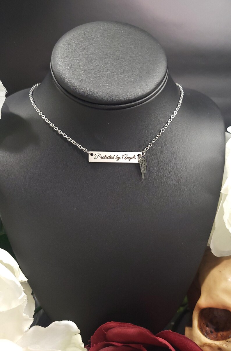 K&ouml;nnte beinhalten: Eine silberfarbene Halskette mit Kette und rechteckigem Anh&auml;nger. Der Anh&auml;nger ist mit den Worten "Protected by Angels" graviert und hat einen kleinen Fl&uuml;gelanh&auml;nger. Die Halskette wird auf einer schwarzen Schaufensterpuppe pr&auml;sentiert.