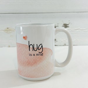 May include: White ceramic mug with a handle, featuring a watercolor design in shades of pink and a small red heart. The mug has the words "hug in a mug" written in black script.