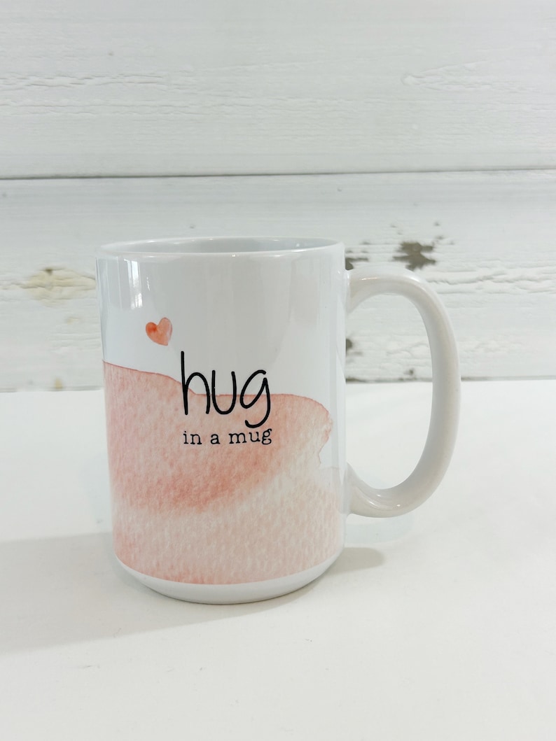 May include: White ceramic mug with a handle, featuring a watercolor design in shades of pink and a small red heart. The mug has the words "hug in a mug" written in black script.