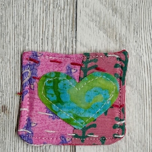 May include: A pink fabric square with a green and blue heart in the center. The heart is outlined in white thread. The fabric is stitched together with white thread.