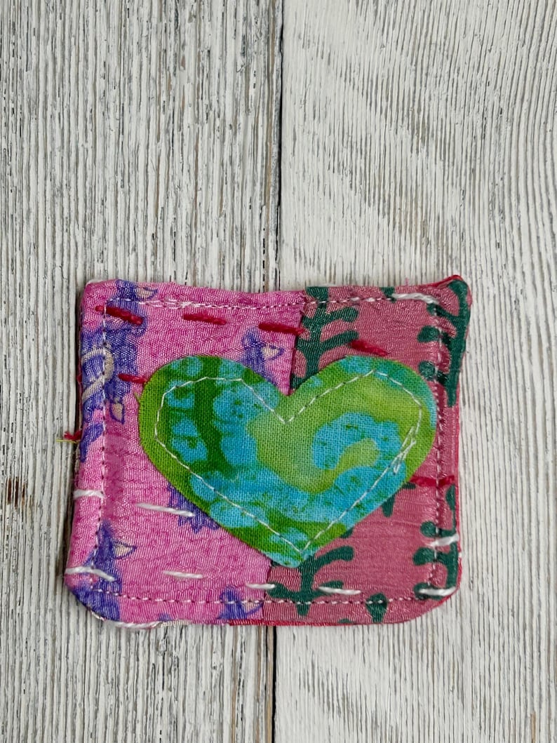 May include: A pink fabric square with a green and blue heart in the center. The heart is outlined in white thread. The fabric is stitched together with white thread.