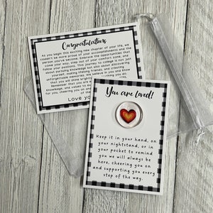 K&ouml;nnte beinhalten: Ein kleiner, klarer, herzf&ouml;rmiger Token mit einem rot-gelben Herzdesign, pr&auml;sentiert auf einer schwarz-wei&szlig; karierten Karte mit der Aufschrift "You are loved!" und "Congratulations." Der Token ist mit einem kleinen, durchsichtigen Beutel verpackt.
