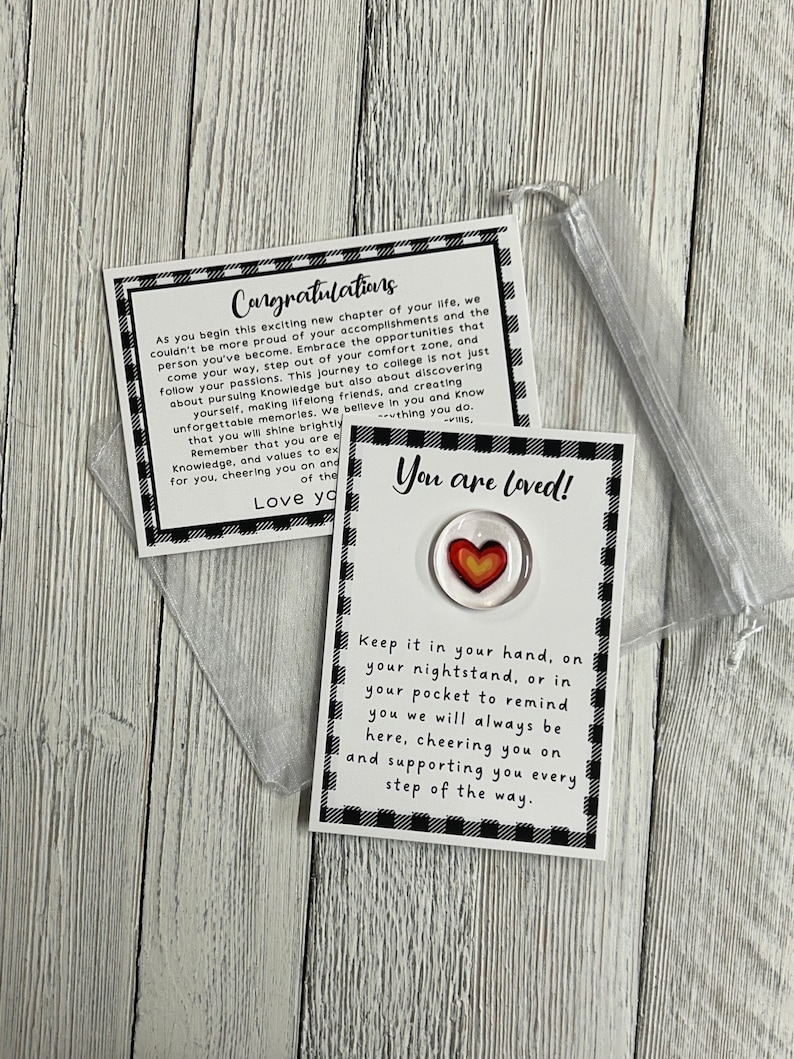 K&ouml;nnte beinhalten: Ein kleiner, klarer, herzf&ouml;rmiger Token mit einem rot-gelben Herzdesign, pr&auml;sentiert auf einer schwarz-wei&szlig; karierten Karte mit der Aufschrift "You are loved!" und "Congratulations." Der Token ist mit einem kleinen, durchsichtigen Beutel verpackt.