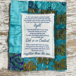 May include: A small, colorful fabric square with a white label containing a poem about faith and hope. The poem reads: "In this quilt square, small and bright, are prayers for you, both day and night For strength and comfort, peace and light, To lift your heart, and make burdens light. Hold this little quilt in your hand, When life throws you those things you did not plan. To calm your heart, mind, and soul, as a reminder that God is in Control. May it catch each tear you weep, And give it to God and go to sleep. Know in your heart, with every seam, His love for you is mightier than the sea."
