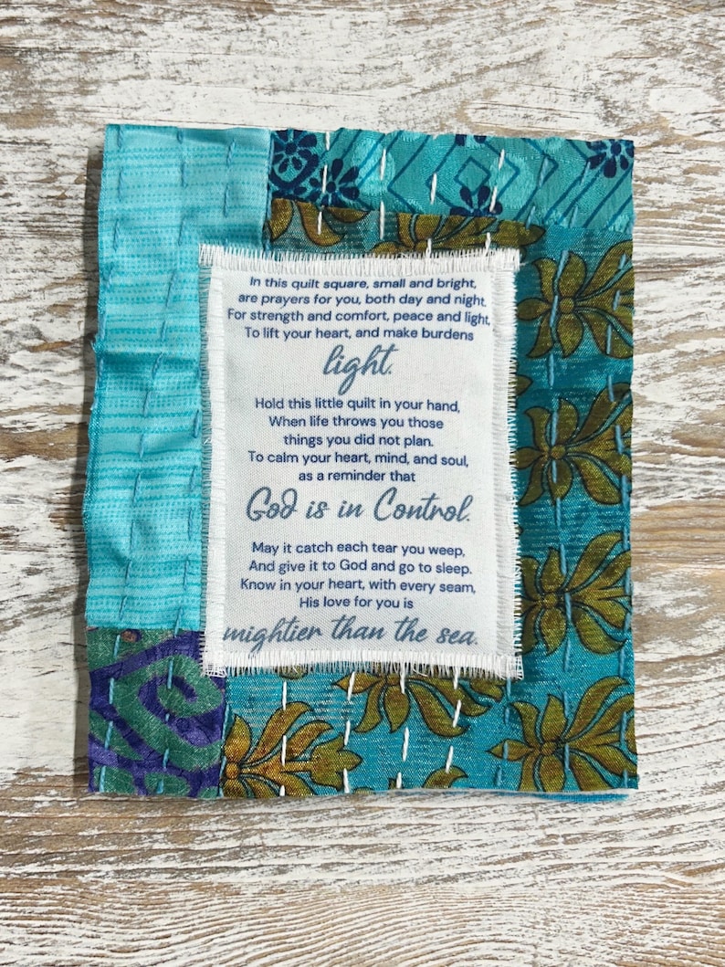 May include: A small, colorful fabric square with a white label containing a poem about faith and hope. The poem reads: "In this quilt square, small and bright, are prayers for you, both day and night For strength and comfort, peace and light, To lift your heart, and make burdens light. Hold this little quilt in your hand, When life throws you those things you did not plan. To calm your heart, mind, and soul, as a reminder that God is in Control. May it catch each tear you weep, And give it to God and go to sleep. Know in your heart, with every seam, His love for you is mightier than the sea."